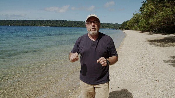 Man in a hat and glasses stands on a beach by the ocean