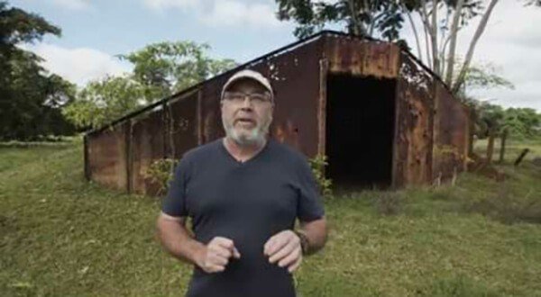 Man in glasses stands in front of a rusty corrugated metal structure.