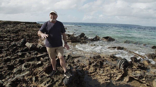 Man standing on rocky shore with wreckage near the ocean