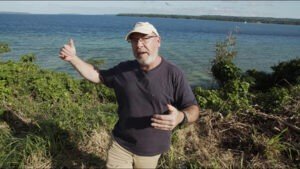 Man in a cap gestures towards a blue lake, discussing videos.