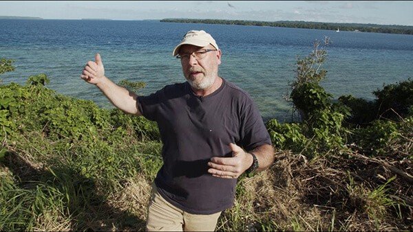 Man in a cap gestures towards a blue lake, discussing videos.