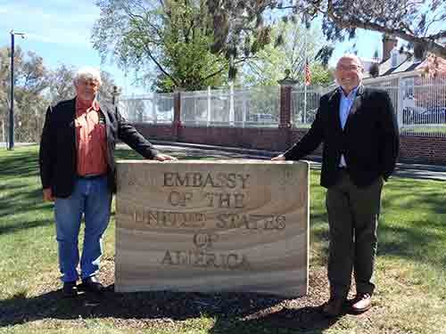 Two men stand beside a stone monument that reads "Embassy of the United States of America" in Canberra.