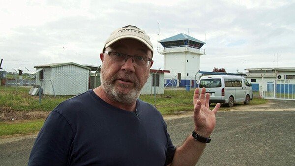 Man in cap and glasses gestures near an airport control tower and buildings.