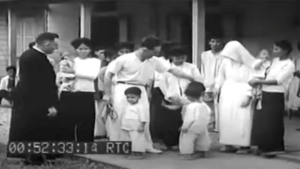 Priest with children and adults in a video still, 00:52:33:14 timestamp