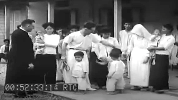 Priest with children and adults in a video still, 00:52:33:14 timestamp