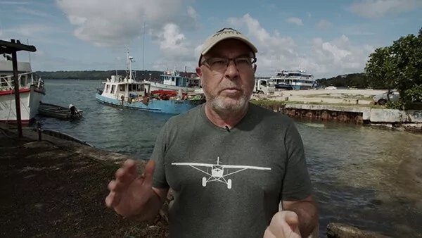 Man in a t-shirt with a small plane graphic stands in a harbor with boats.