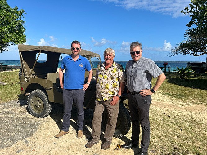 Three men stand next to a vintage military jeep on a sunny beach.