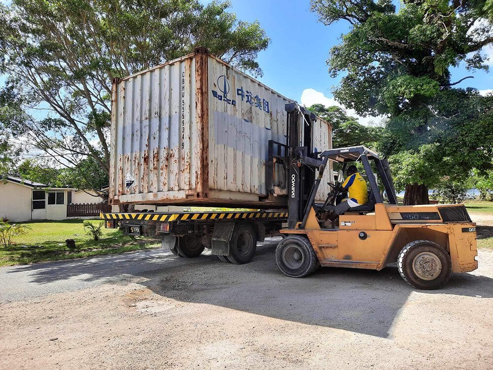 Forklift loading a rusty shipping container onto a truck in a tropical setting.
